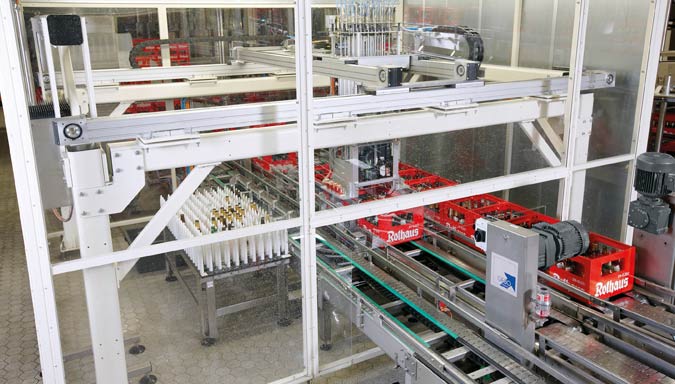 The robot cell with overhead portal is supplied with empties from two parallel roller conveyors.